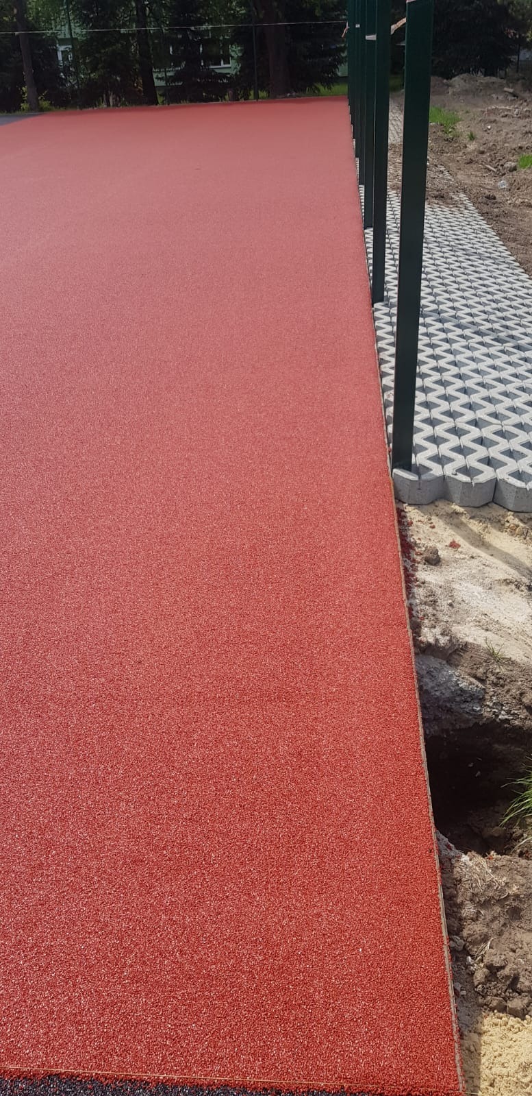 Czerwona, gumowa nawierzchnia boiska sportowego przylegająca do ażurowych, betonowych płyt chodnikowych i zielonych słupków ogrodzenia.