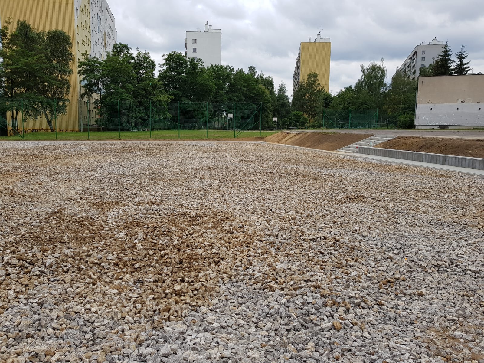 Przygotowany teren pod budowę, z wysypanym tłuczniem, widoczne schody i krawężniki, w tle bloki mieszkalne.
