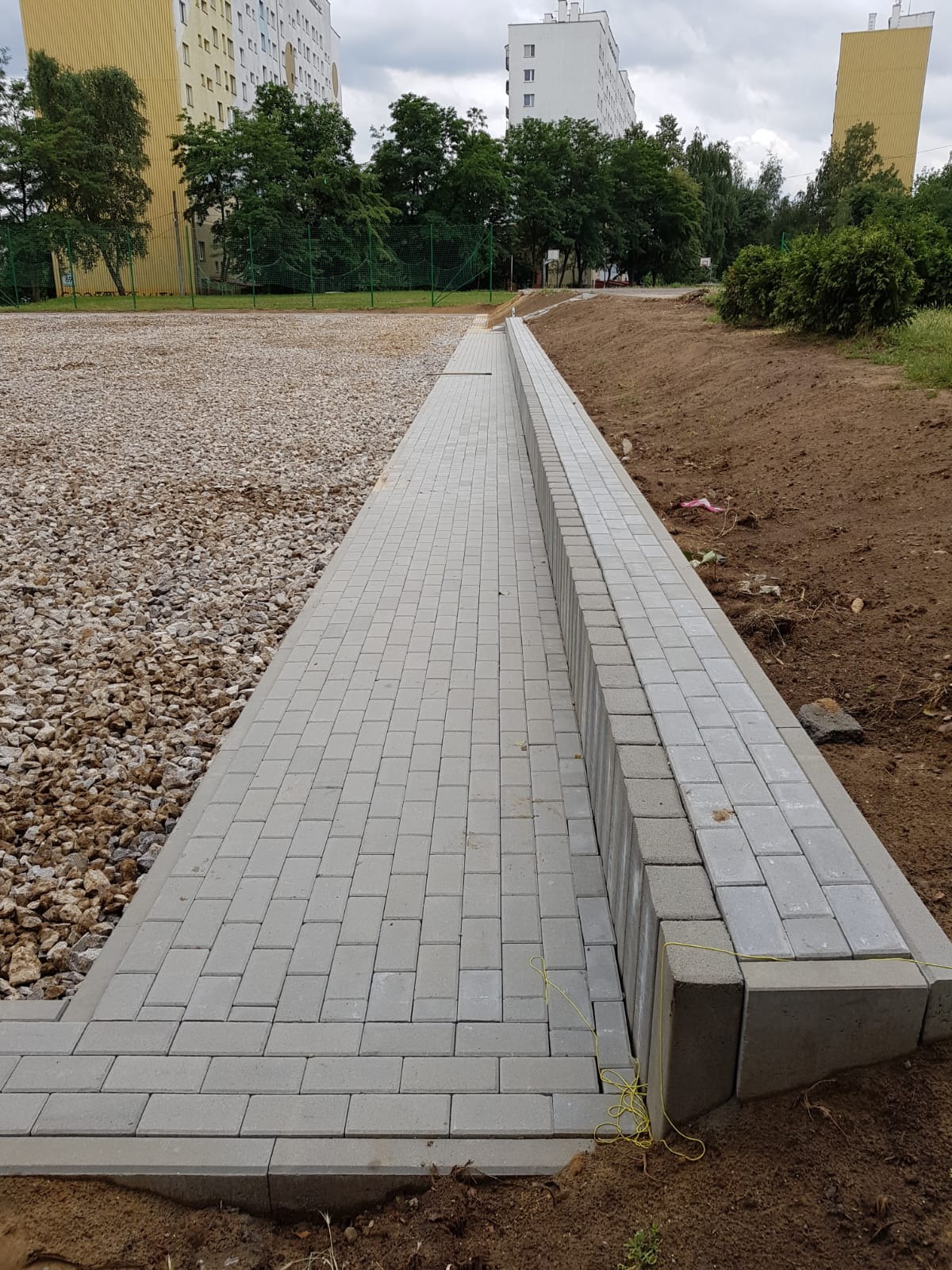 Świeżo ułożona ścieżka z szarej kostki brukowej prowadząca wzdłuż gruntu przygotowanego pod trawnik, obok placu wysypanego żwirem, na tle bloków mieszkalnych w pochmurny dzień.