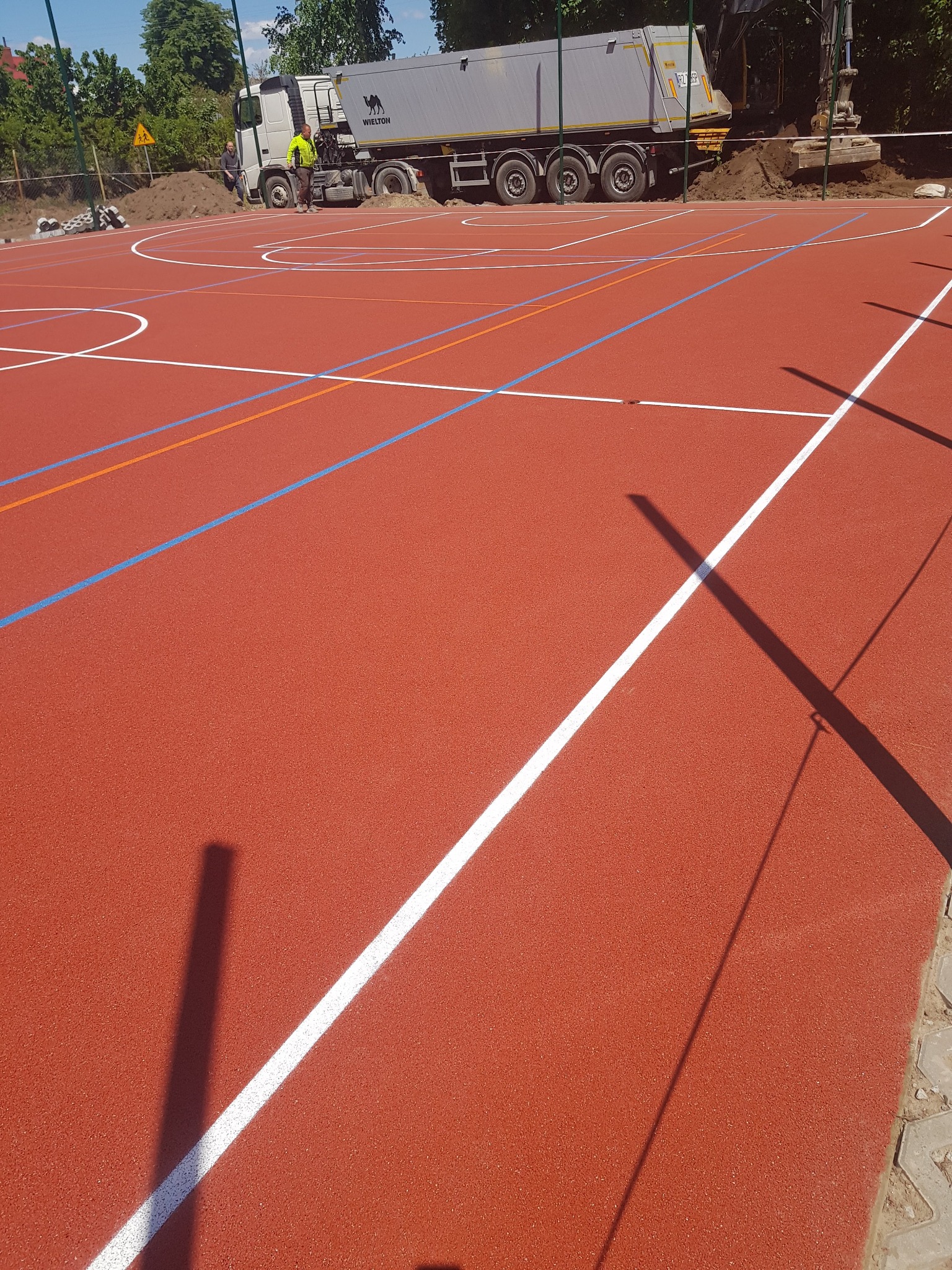 Nowa, czerwona nawierzchnia boiska sportowego z wyraźnie oznaczonymi liniami w kolorach białym, niebieskim i pomarańczowym. W tle, ciężarówka wyładowuje ziemię, a w pobliżu pracuje operator koparki.