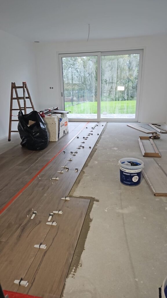 Układanie podłogi z płytek imitujących drewno w jasnym wnętrzu. Widoczne narzędzia, klej i poziomice. Praca w toku, z widokiem na okno tarasowe i ogród.