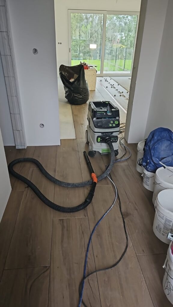 Układanie drewnopodobnych paneli podłogowych w jasnym wnętrzu. Widoczny odkurzacz przemysłowy Festool, wiadro z farbą i czarny worek na śmieci. Duże okno w tle.