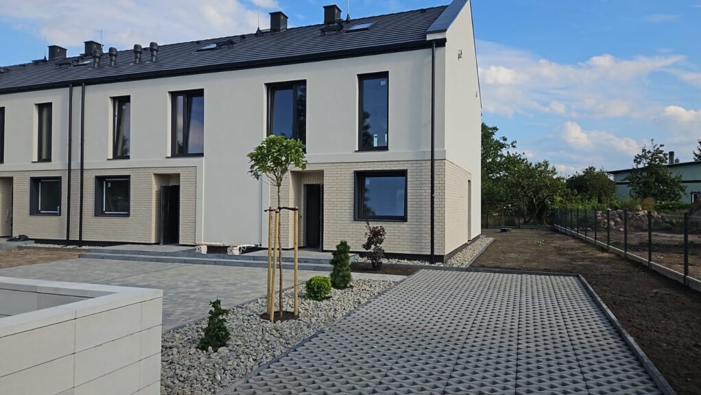 Nowoczesne, szeregowe domy z jasną elewacją i ciemnymi oknami, z zagospodarowanym terenem: kostka ażurowa, kamień ozdobny i młode drzewka. Ujęcie w słoneczny dzień.