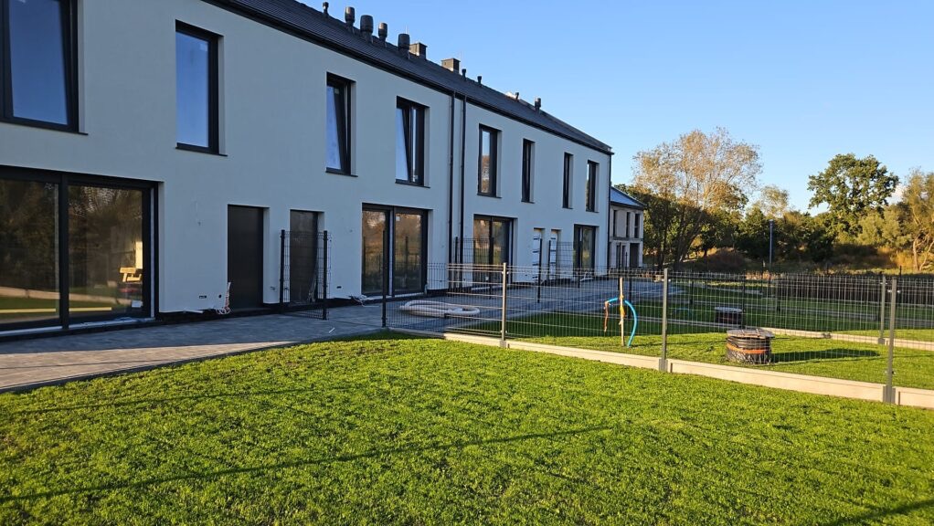 Nowoczesny szeregowiec z białymi ścianami i czarnymi oknami, widok z ogrodu z trawnikiem i ogrodzeniem. Częściowo wykończony teren wokół budynku.