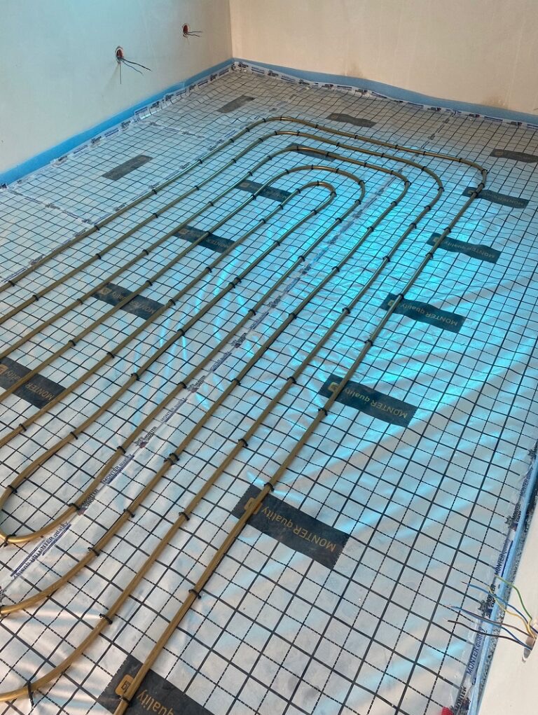 Ułożone rury ogrzewania podłogowego na folii z nadrukiem siatki, widoczne kable elektryczne i fragment ściany z puszkami. Instalacja przed wylaniem posadzki.