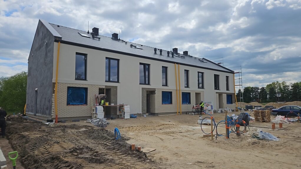 Budowa szeregowca: elewacja częściowo otynkowana, okna wstawione, widoczne rusztowania i materiały budowlane. Prace w toku, teren wokół budynku przygotowany.