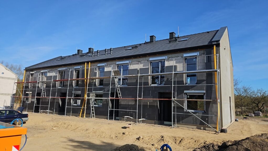 Budynek w trakcie ocieplania, widoczne rusztowania, okna z roletami, czarna izolacja termiczna na ścianach. Częściowo otynkowana ściana szczytowa.