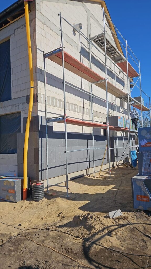 Budowa domu z białych bloczków, ocieplonego szarym styropianem. Widoczne rusztowania i materiały budowlane. Żółta rura spustowa wzdłuż ściany.