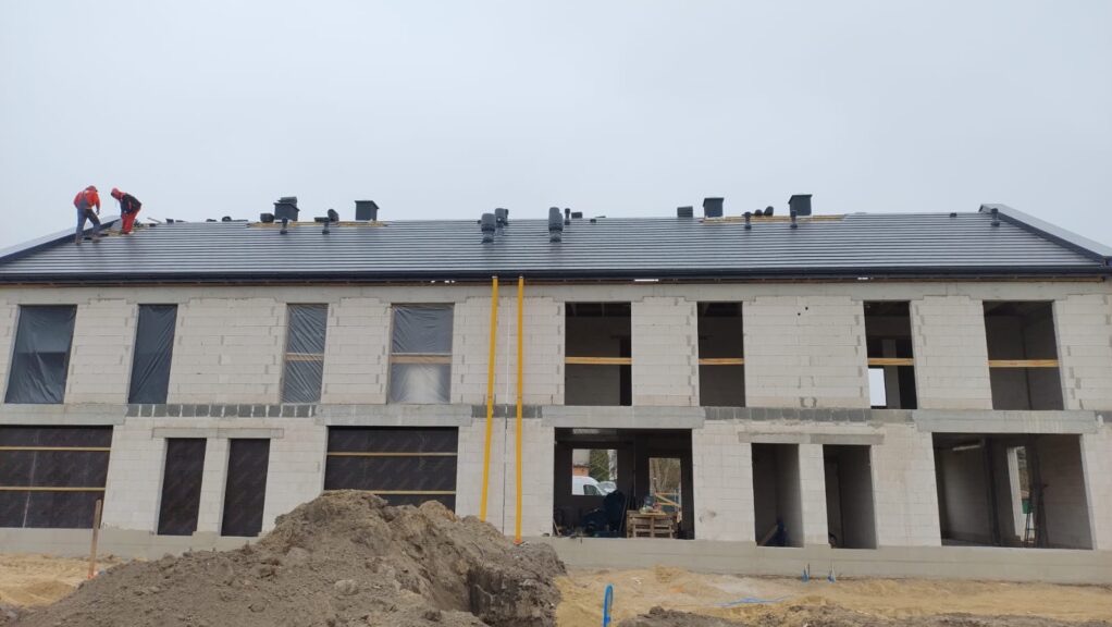 Wznoszony budynek z białego bloczka, z czarnym dachem. Na dachu dwóch robotników w czerwonych kombinezonach. Okna zabezpieczone folią, widoczna sterta ziemi.