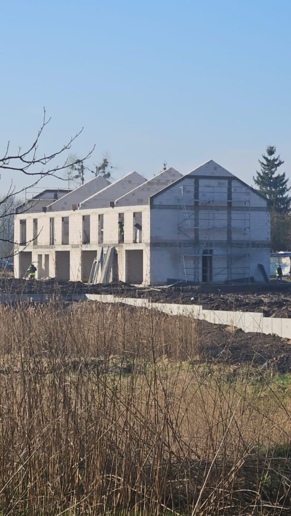 Budowa szeregowców z białego bloczka, widoczne rusztowania i fundamenty. Na pierwszym planie wysoka trawa, w tle błękitne niebo i drzewa.