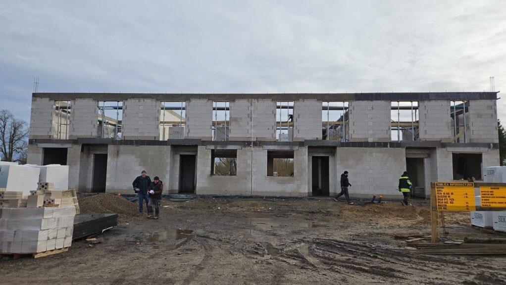 Surowy stan bliźniaka z betonu komórkowego w trakcie budowy. Widoczne niezadaszone otwory okienne i drzwiowe. Na pierwszym planie materiały budowlane i ekipa budowlana.