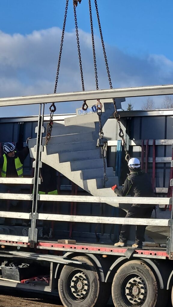 Transport prefabrykowanych, betonowych schodów na budowę. Dźwig z łańcuchami podnosi element. Pracownicy w kaskach i kamizelkach asekurują ładunek na ciężarówce.