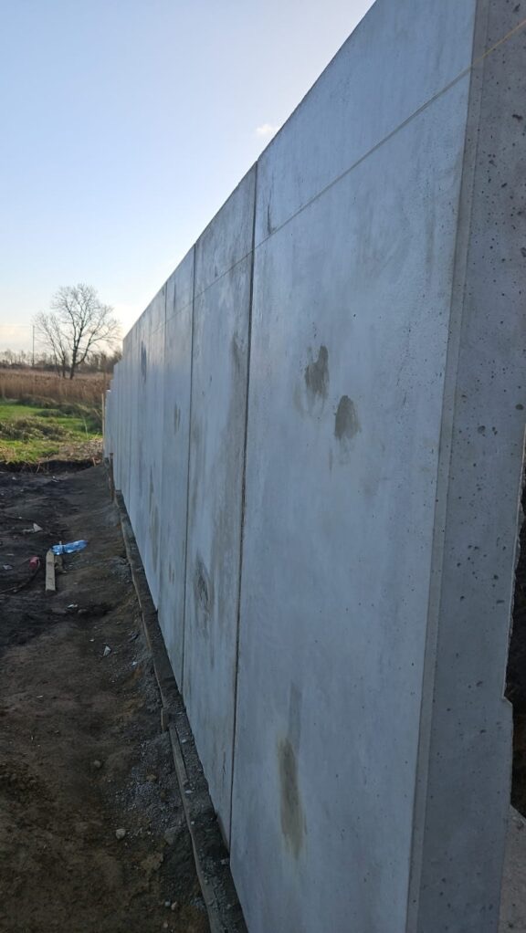 Szara, betonowa ściana oporowa z widocznymi łączeniami paneli, na tle ziemi i zieleni. Ujęcie z dołu, podkreślające wysokość konstrukcji.