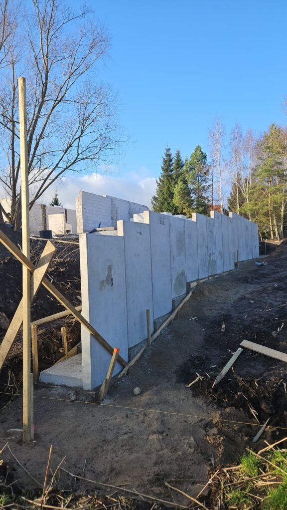 Wznoszenie betonowej ściany oporowej na nierównym terenie, widoczne drewniane podpory i fragmenty fundamentów. W tle drzewa i błękitne niebo.