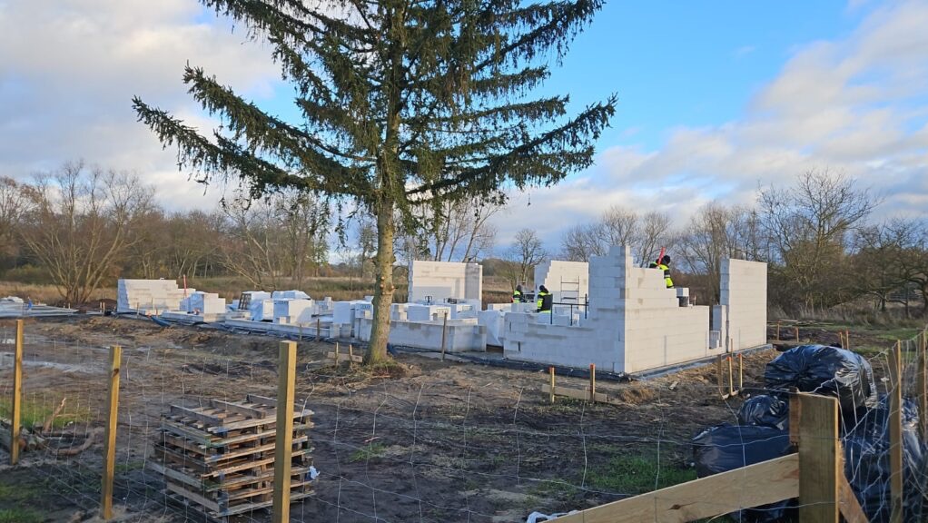 Wznoszenie ścian domu z bloczków betonu komórkowego na działce, widoczni pracownicy w odblaskowych kamizelkach, w tle drzewa i błękitne niebo.
