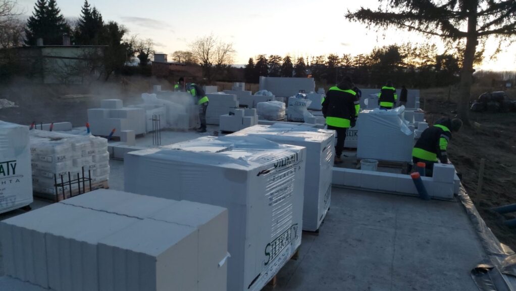 Wznoszenie ścian z bloczków Ytong Silka na fundamencie. Widoczni pracownicy w odblaskowych kamizelkach. Część bloczków w oryginalnych opakowaniach.