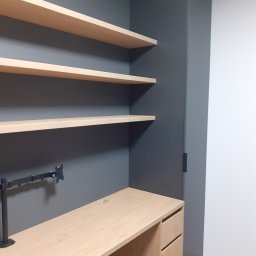 MARCIN KOŁODZIEJCZAK - Biurko z trzema szufladami, jasne półki wiszące na szarej ścianie, ramię uchwytu monitora, minimalistyczne wnętrze.