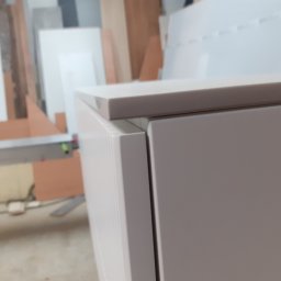 MARCIN KOŁODZIEJCZAK - Fragment szafki z białymi frontami i blatem, widoczne detale łączenia elementów, w tle warsztat stolarski z płytami meblowymi.