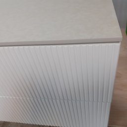 MARCIN KOŁODZIEJCZAK - Fragment szafki z białymi, pionowo żłobionymi frontami i jasnym blatem, ustawionej na płycie drewnianej.