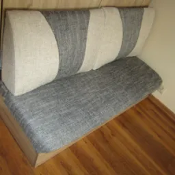 Narożna ławka tapicerowana szarą tkaniną z poduszkami w beżowo-szare pasy, z drewnianą podstawą i oparciem, na tle drewnianej podłogi i ściany w odcieniu écru.