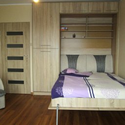 MARCIN KOŁODZIEJCZAK - Wielofunkcyjny mebel: łóżko chowane w szafie z półkami, obok drzwi z przeszkleniami i fotel z tapicerką w odcieniach szarości.