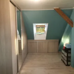 Adaptacja poddasza ze skośnym oknem dachowym i zabudową szaf z przesuwnymi drzwiami w jasnym odcieniu drewna.