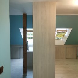 MARCIN KOŁODZIEJCZAK - Wnętrze poddasza z drewnianą kolumną, jasnymi panelami podłogowymi i zabudową pod skosami, widok na okno dachowe.