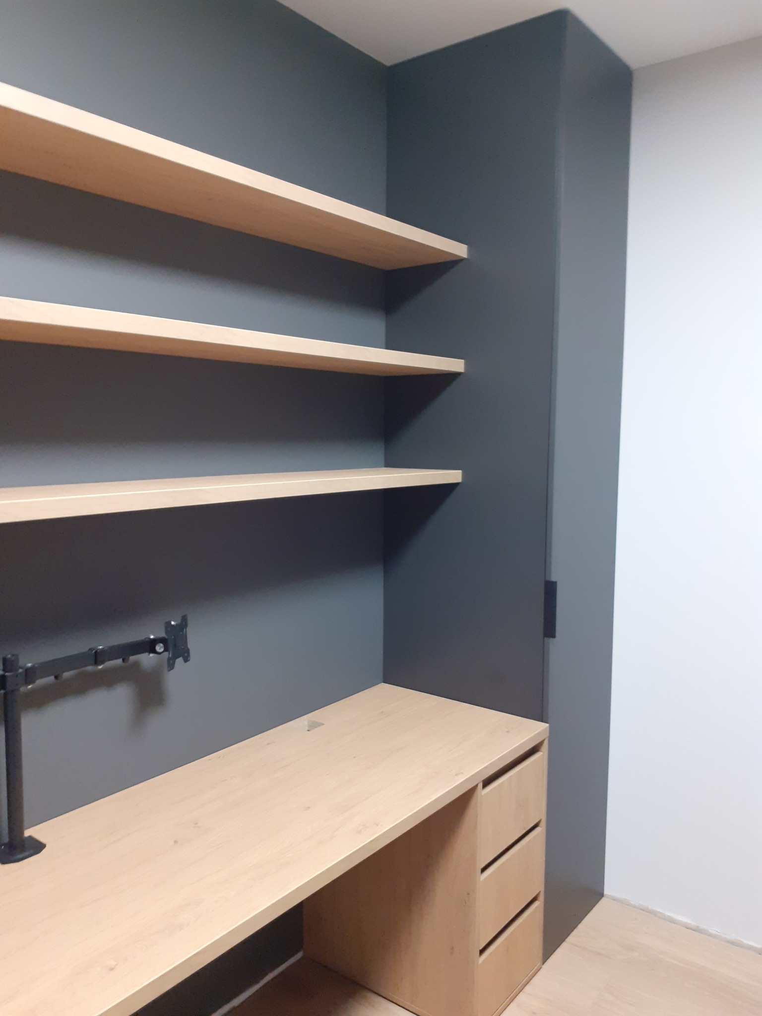 Biurko z trzema szufladami, jasne półki wiszące na szarej ścianie, ramię uchwytu monitora, minimalistyczne wnętrze.