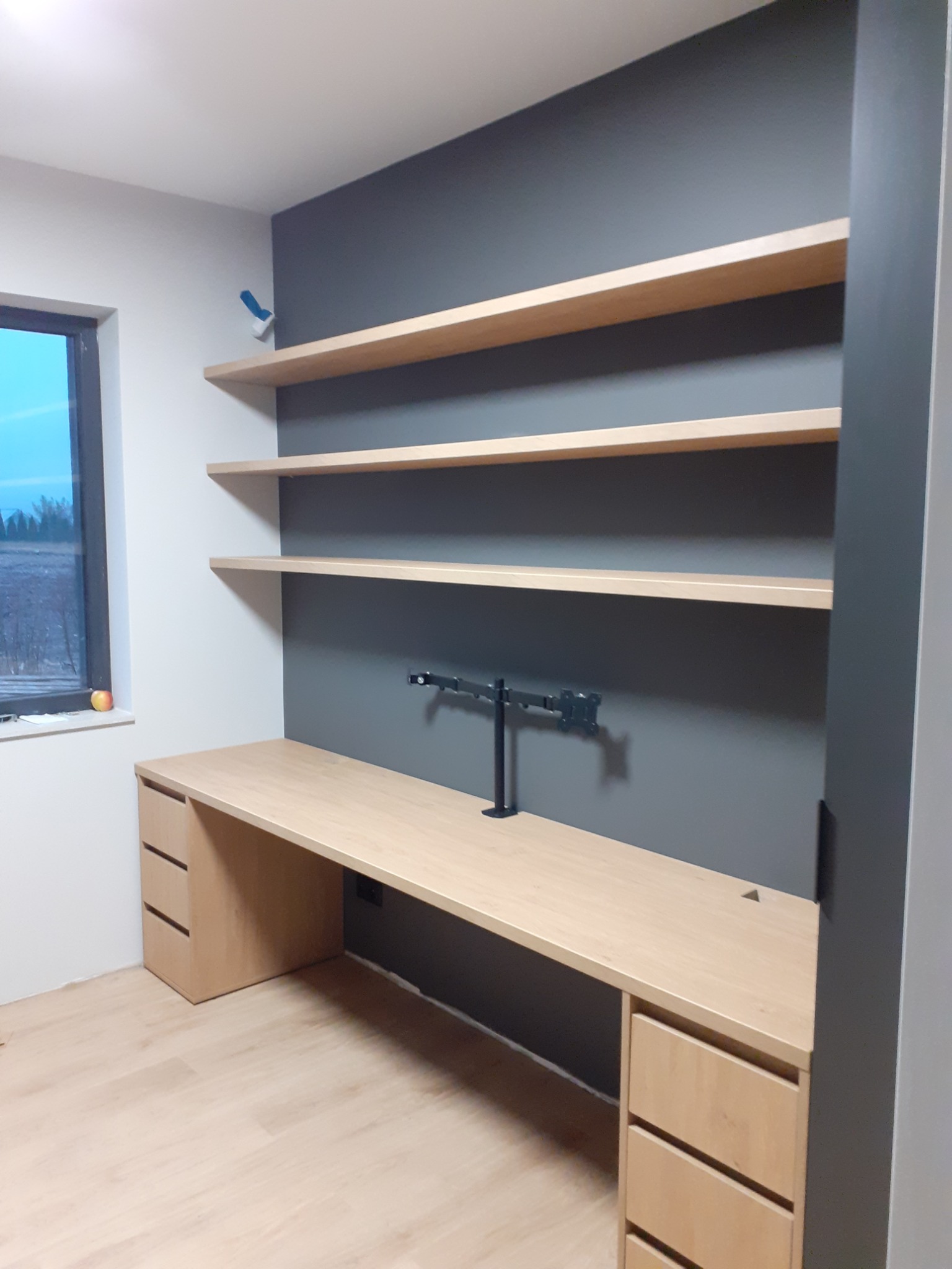 Jasne biurko z szufladami i półkami na szarej ścianie, z zamontowanym uchwytem na monitor, widok na zewnątrz przez okno z jabłkiem na parapecie, minimalistyczny design.