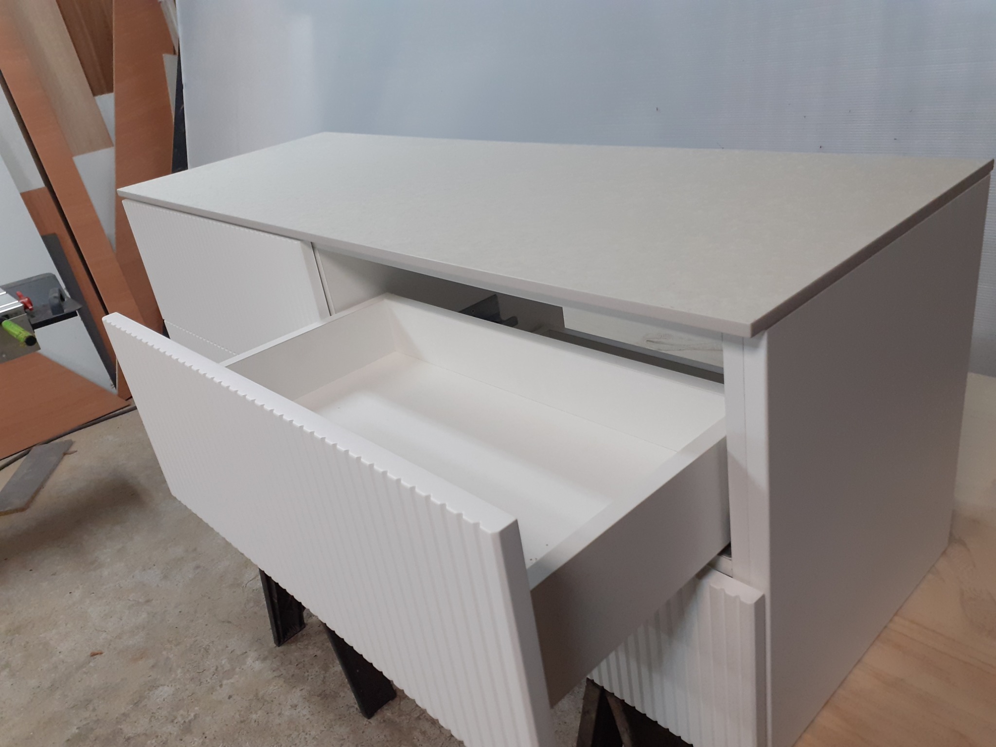 Biała szafka z szufladami o ryflowanych frontach, otwarta szuflada ukazuje wnętrze, widoczne elementy warsztatu w tle.