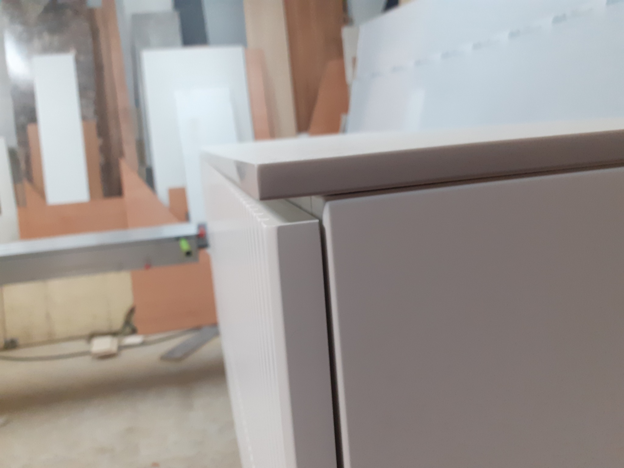 Fragment szafki z białymi frontami i blatem, widoczne detale łączenia elementów, w tle warsztat stolarski z płytami meblowymi.