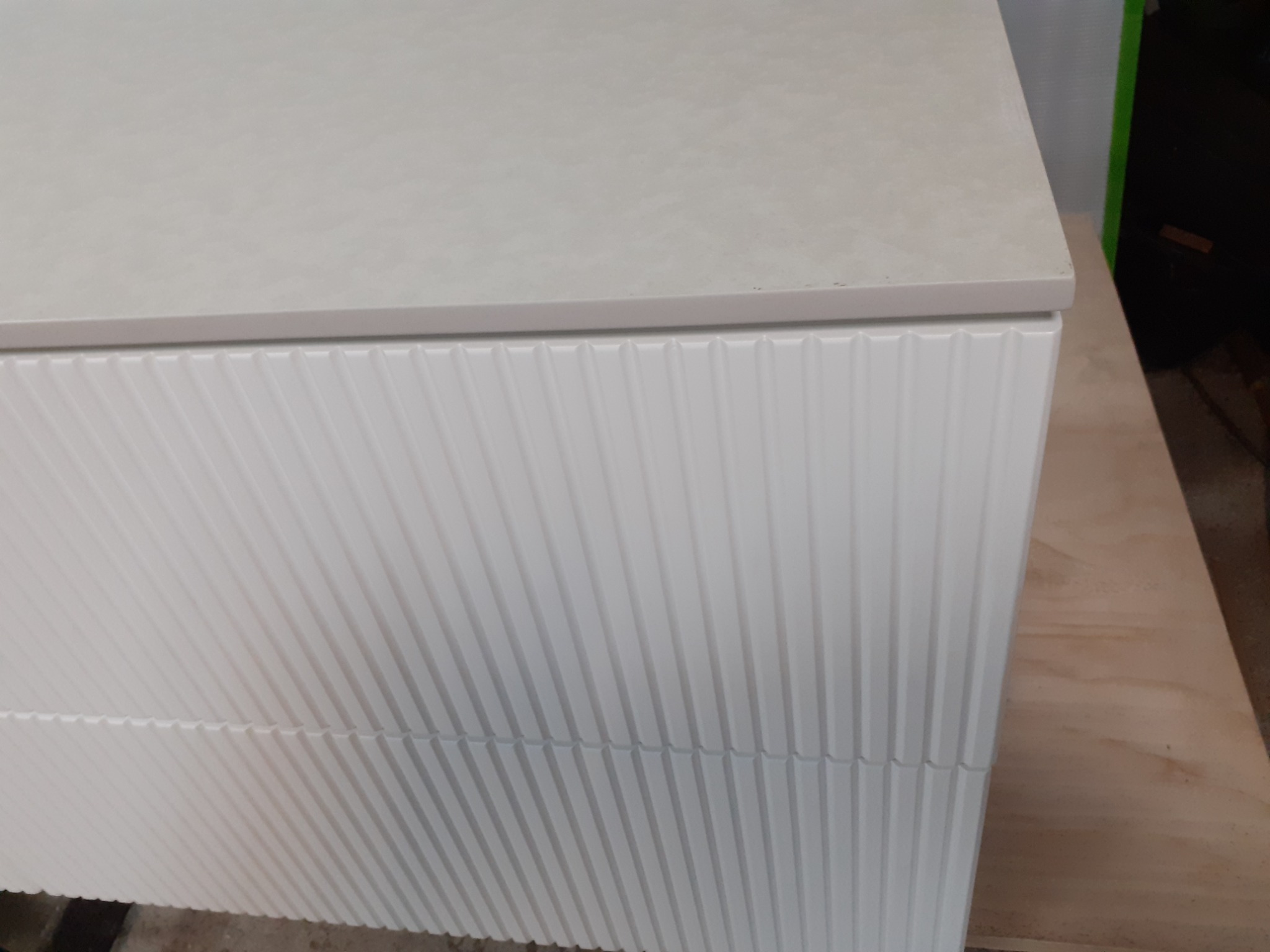 Fragment szafki z białymi, pionowo żłobionymi frontami i jasnym blatem, ustawionej na płycie drewnianej.