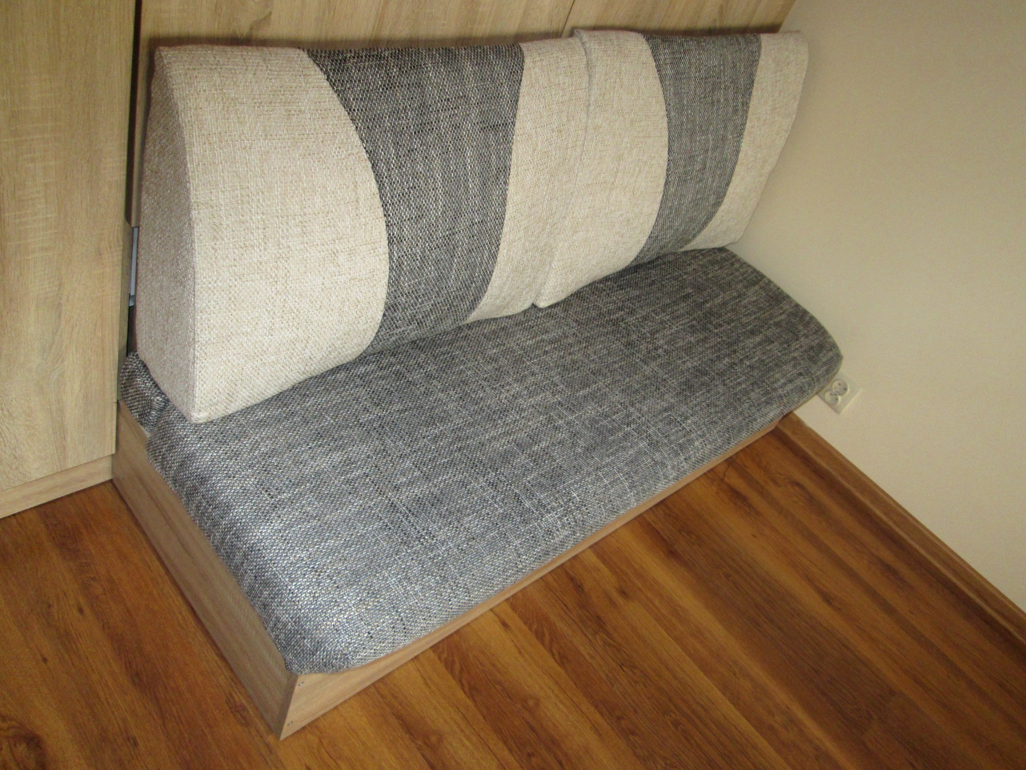 Narożna ławka tapicerowana szarą tkaniną z poduszkami w beżowo-szare pasy, z drewnianą podstawą i oparciem, na tle drewnianej podłogi i ściany w odcieniu écru.