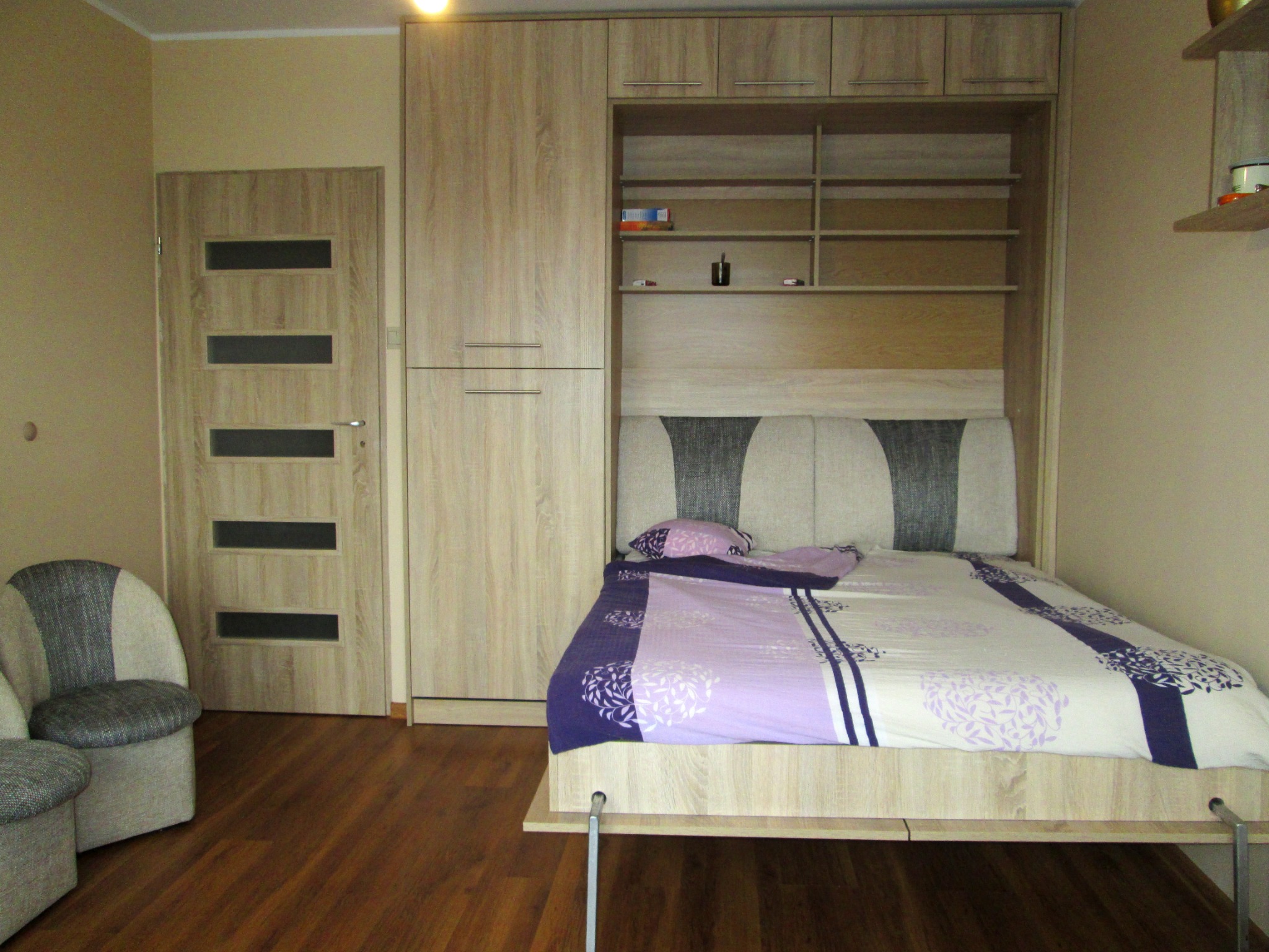 Wielofunkcyjny mebel: łóżko chowane w szafie z półkami, obok drzwi z przeszkleniami i fotel z tapicerką w odcieniach szarości.