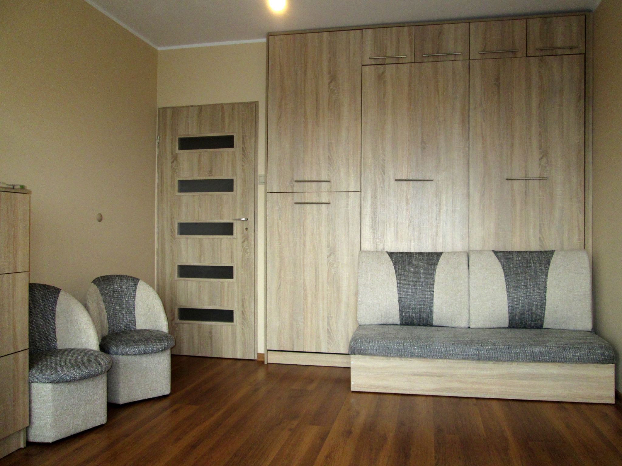 Wnętrze pokoju z drewnianą podłogą, szafą wnękową zintegrowaną z rozkładaną sofą, dwoma krzesłami z szaro-białą tapicerką oraz drzwiami z poziomymi, czarnymi szybami.