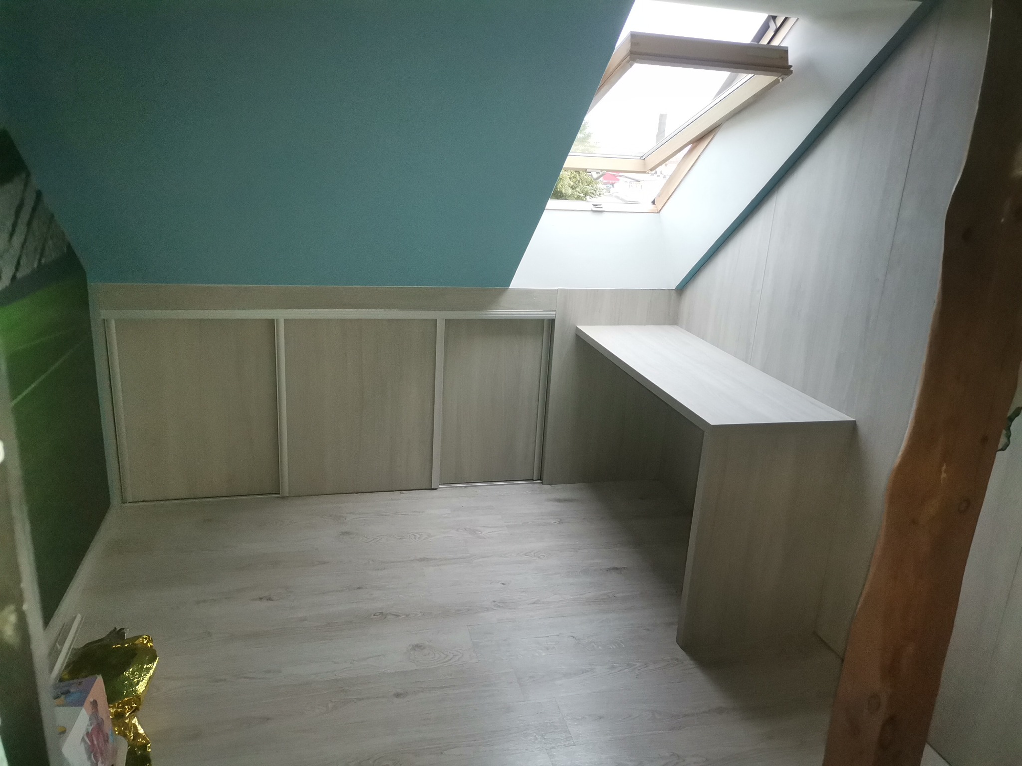 Szafa wnękowa z przesuwnymi drzwiami i biurko zamontowane na poddaszu z oknem dachowym, szare panele na podłodze, niebieski sufit.