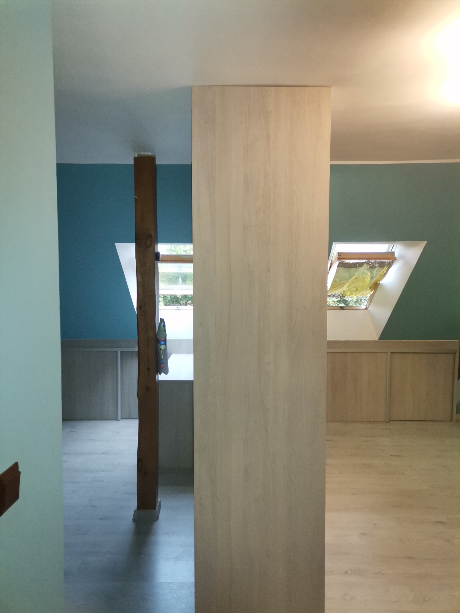 Wnętrze poddasza z drewnianą kolumną, jasnymi panelami podłogowymi i zabudową pod skosami, widok na okno dachowe.