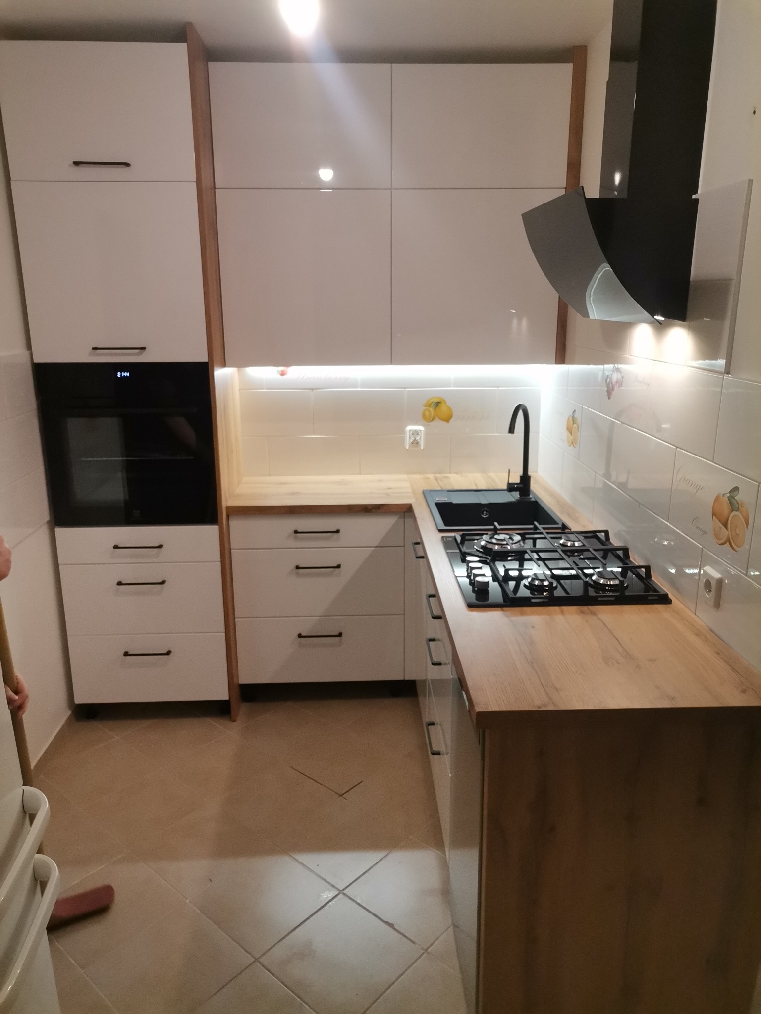 Nowoczesna zabudowa kuchenna w Miejskej Górce: białe fronty szafek z drewnianymi akcentami, czarny piekarnik i płyta gazowa, okap nad płytą, jasne płytki z motywem owocowym na ścianie, drewniany blat.