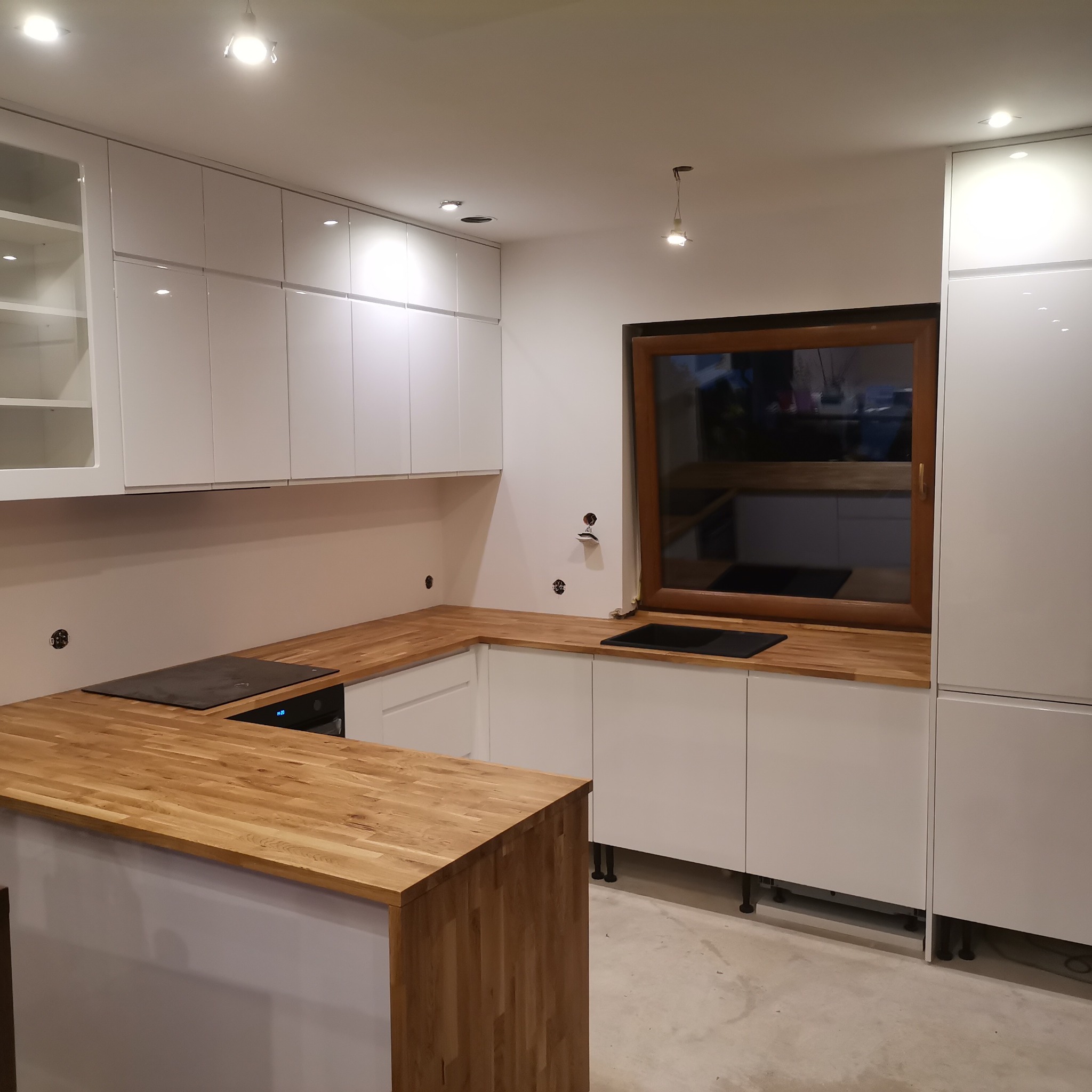 Nowoczesna kuchnia z białymi szafkami i drewnianym blatem, widoczne niedokończone otwory na gniazdka elektryczne.