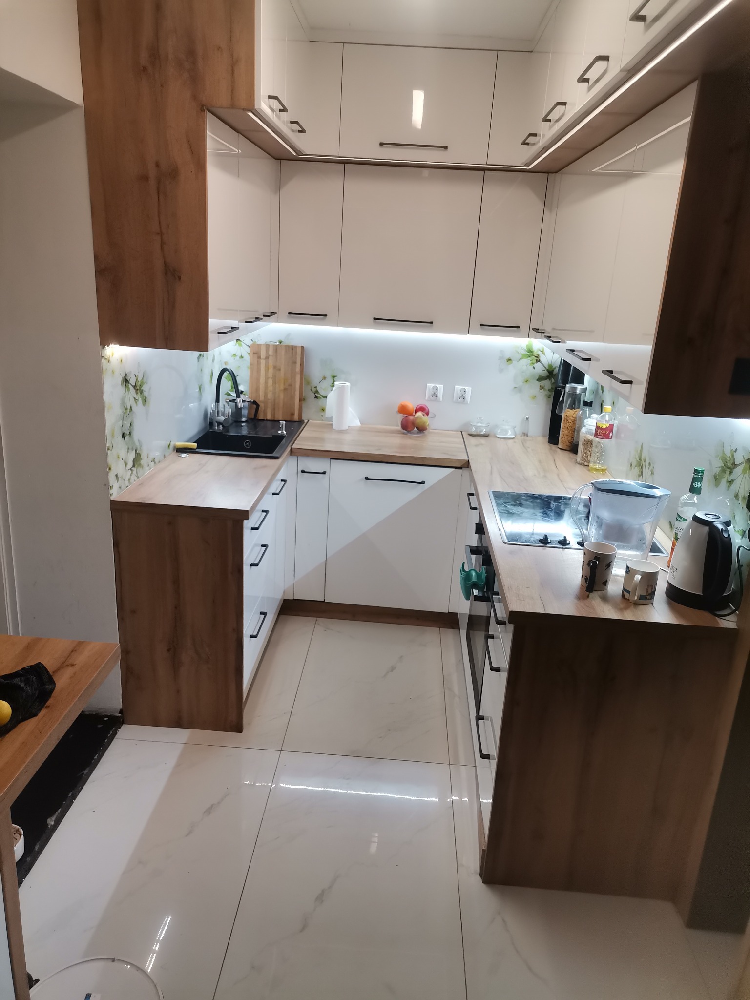 Nowoczesna kuchnia w zabudowie z białymi frontami i drewnianymi akcentami, czarny zlew, marmurowa podłoga, oświetlenie LED pod szafkami.