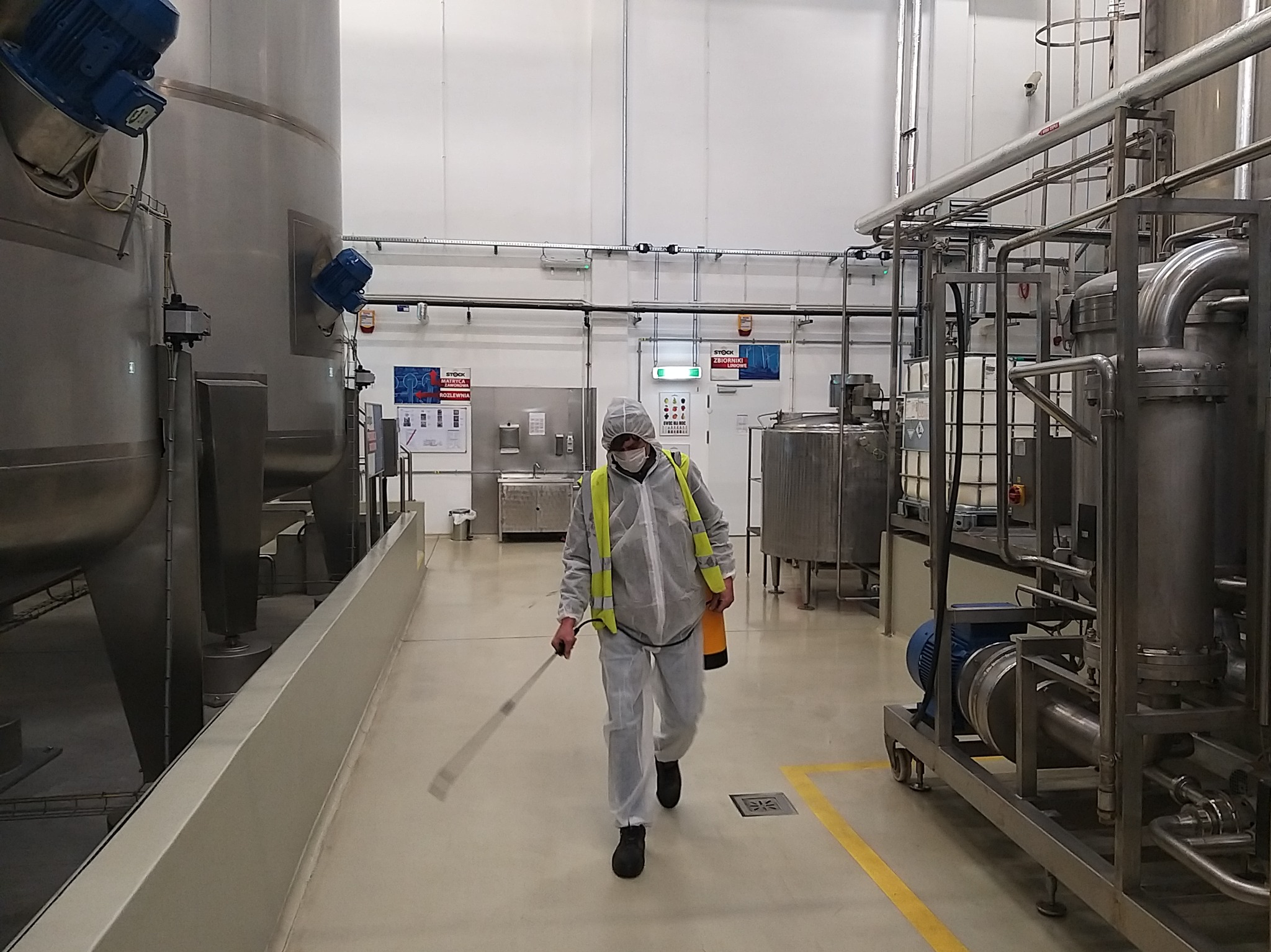 Pracownik w kombinezonie ochronnym i masce dezynfekuje podłogę w hali przemysłowej z dużymi zbiornikami i rurami ze stali nierdzewnej.