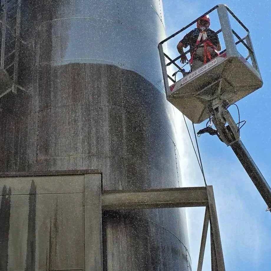 Pracownik w koszu podnośnika czyści ciśnieniowo zewnętrzną powierzchnię przemysłowego zbiornika z szarej stali, widoczne zacieki i rozpryski wody.