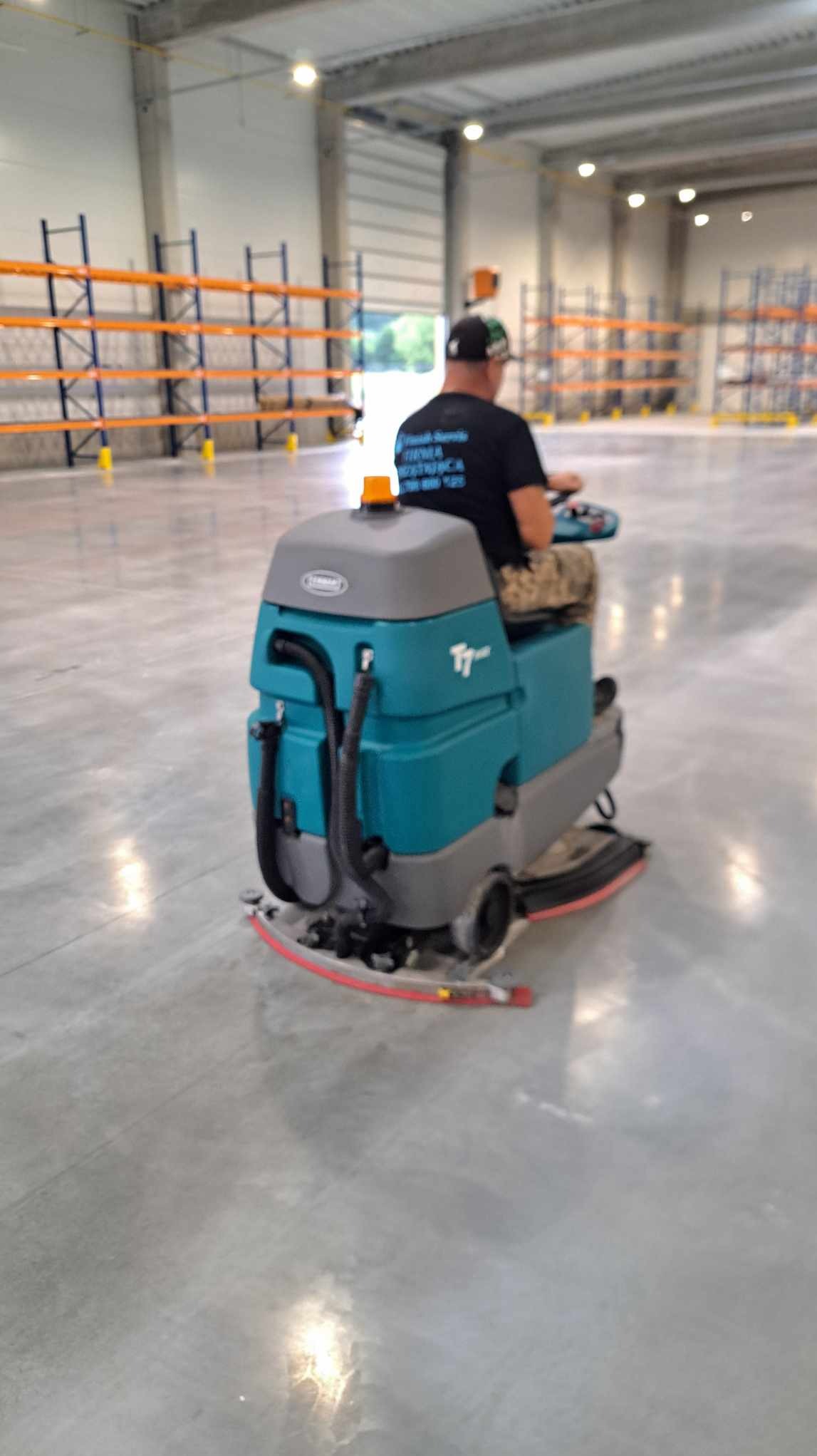 Operator na pojeździe czyszczącym T7 marki Tennant, poleruje betonową posadzkę w hali magazynowej z pomarańczowymi regałami w tle.