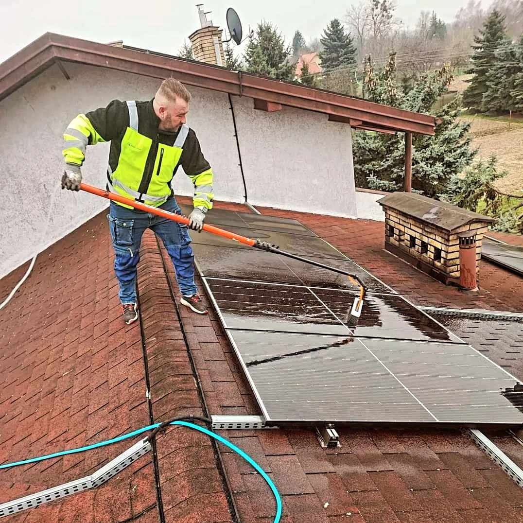 Mężczyzna w odblaskowej kurtce czyści mokre panele słoneczne na dachu domu za pomocą szczotki na długim kiju, widoczne kable i otoczenie z drzewami.
