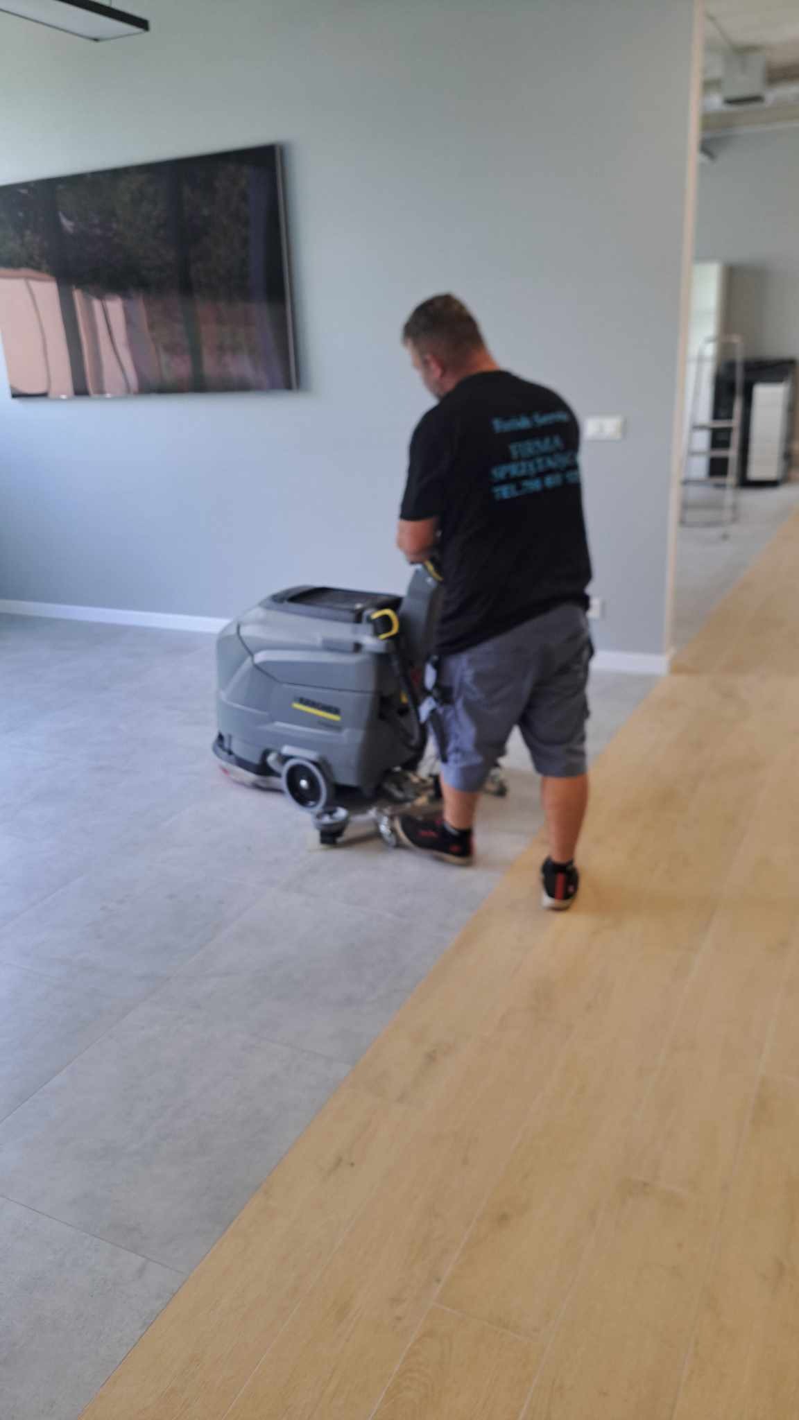 Mężczyzna w czarnej koszulce z napisem firmy sprzątającej obsługuje szaro-żółtą maszynę czyszczącą podłogę w pomieszczeniu z dwoma rodzajami podłóg: szare płytki i panele drewnopodobne.