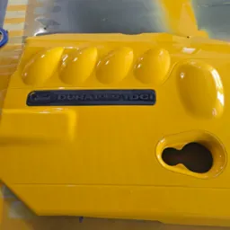 Żółta, błyszcząca pokrywa silnika Ford Duratorq TDCI po lakierowaniu, leżąca na powierzchni roboczej z taśmą maskującą. Widoczne refleksy światła.