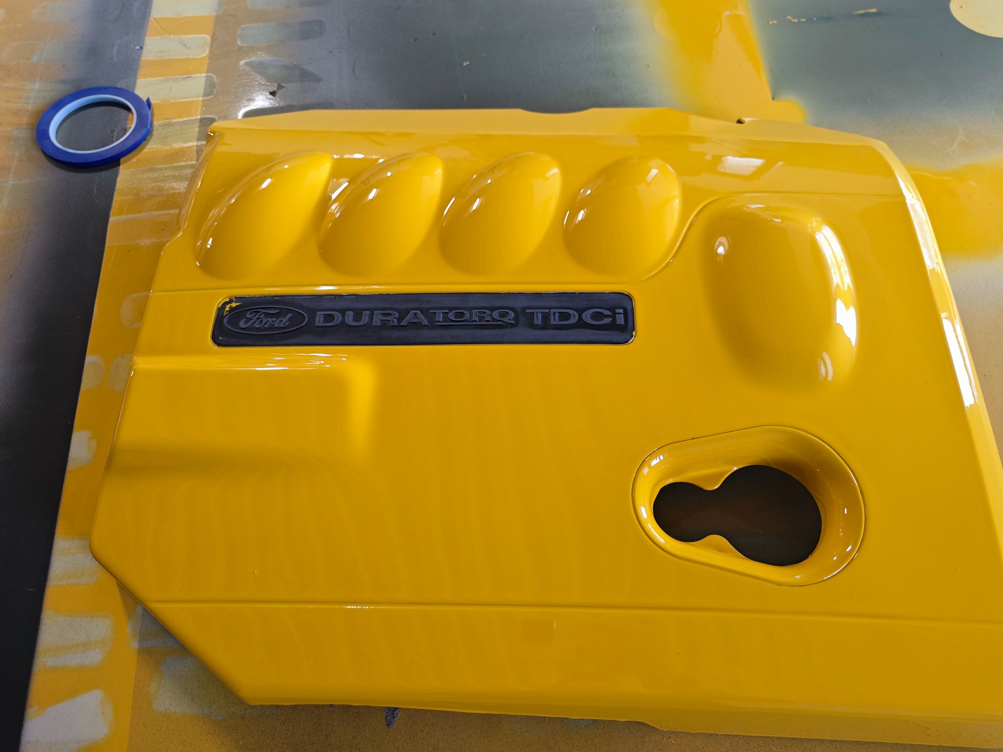 Żółta, błyszcząca pokrywa silnika Ford Duratorq TDCI po lakierowaniu, leżąca na powierzchni roboczej z taśmą maskującą. Widoczne refleksy światła.