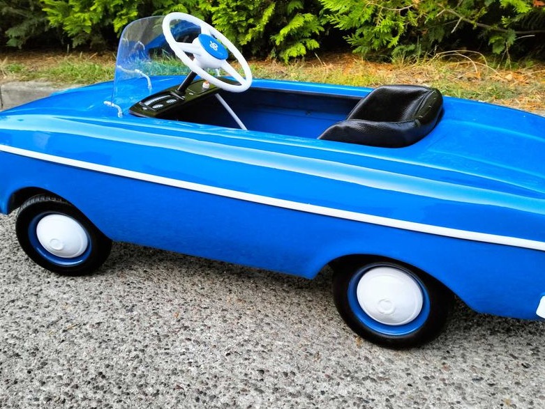 Niebieski, odrestaurowany samochodzik-zabawka z białymi akcentami, widoczny na zewnątrz, na tle zieleni. Błyszczące wykończenie, detale retro.