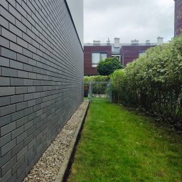 Pan Ogrodnik - Widok z boku domu na trawnik z krzewami i metalową furtką, wąski pas drobnych kamieni przy ścianie budynku.