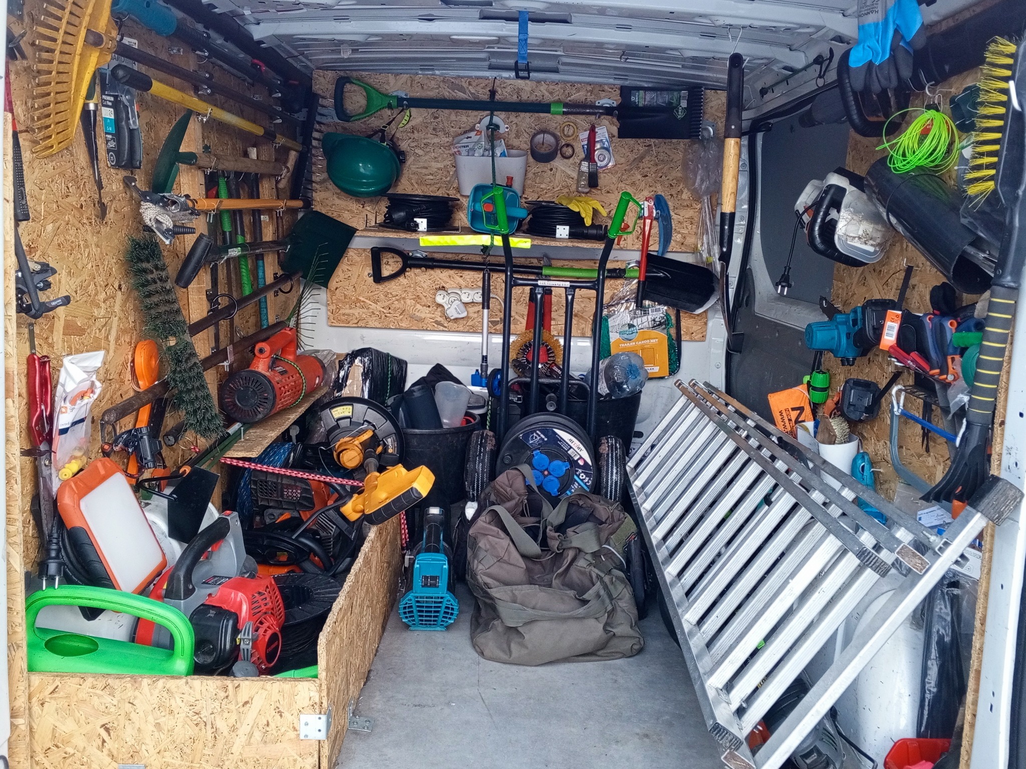 Wnętrze furgonetki wypełnionej narzędziami ogrodniczymi: kosiarki, piły, grabie, łopaty, drabina. Sprzęt uporządkowany na ścianach i podłodze, gotowy do pracy.